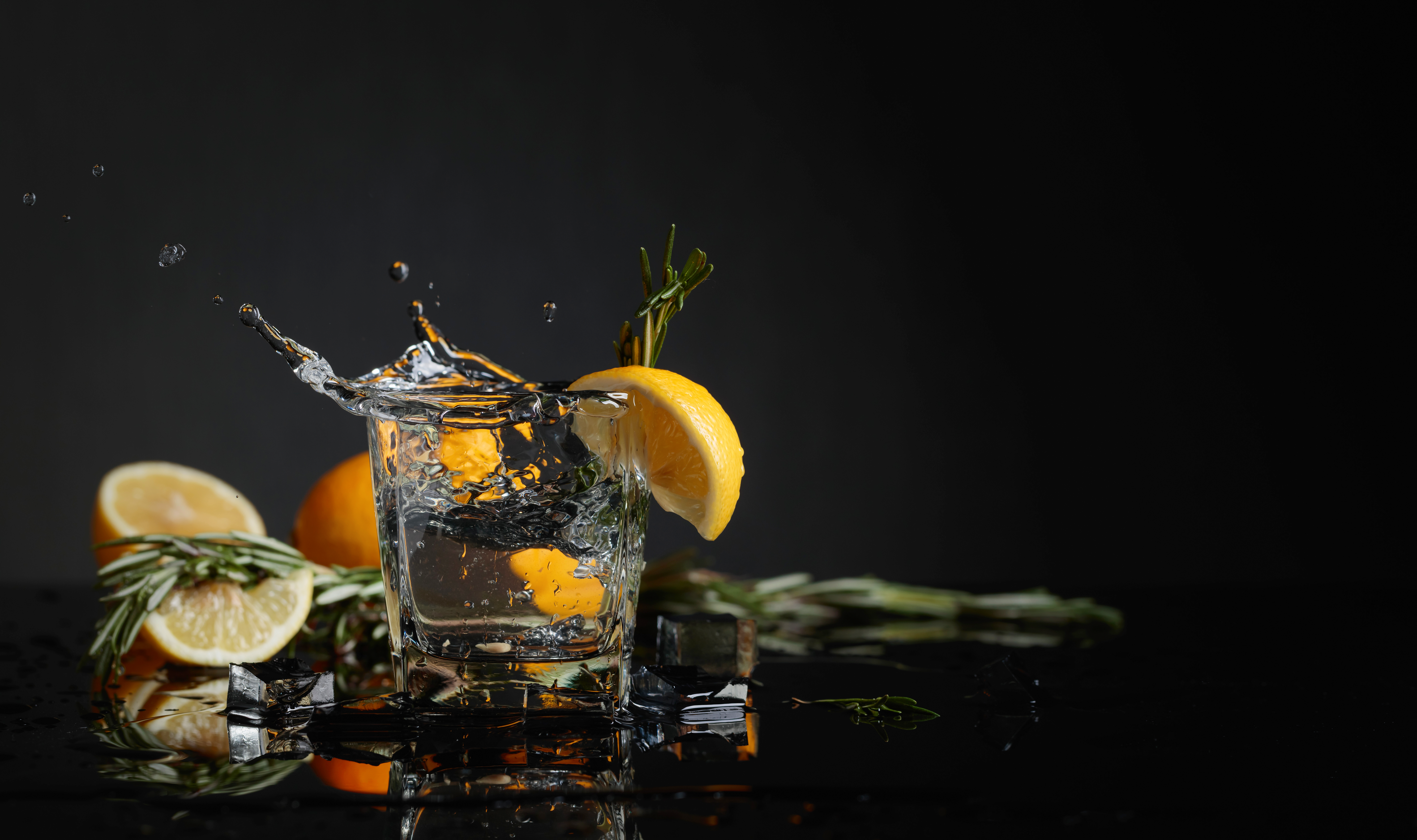 Cocktail gin-tonic with lemon slices and twigs of rosemary. Piece of ice falls into the glass. Copy space.